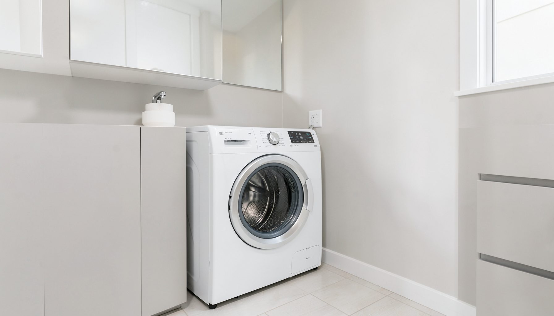 A clean modern laundry room with a front-loading washing machine, soft neutral tones of white and pale gray, calm and tidy